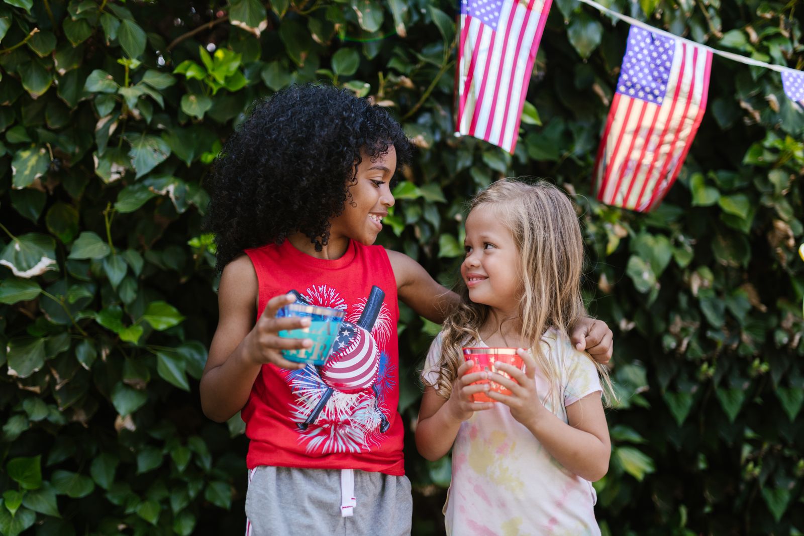 Two smiling children holding cups under American flag bunting in a garden